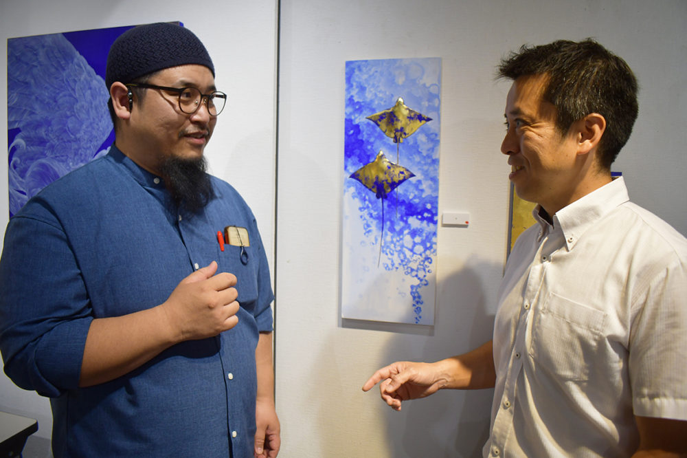 （画像３アイキャッチ兼用：個展会場で談笑する画家の小笠原弘樹さん（左）と小笠原一生准教授。後ろの作品は青地に金箔（きんぱく）で装飾された２匹のエイを描いた「道」）